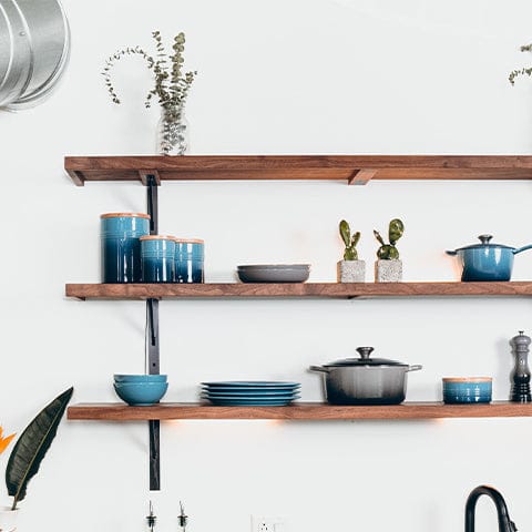 3 long kitchen shelves hanging against a white wall. Each shelf has blue dishes on them with small accents of gray and green from plants.