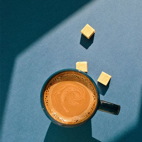 A cup of coffee sitting on a blue table with 3 cubes of sugar tossed next to it. 
