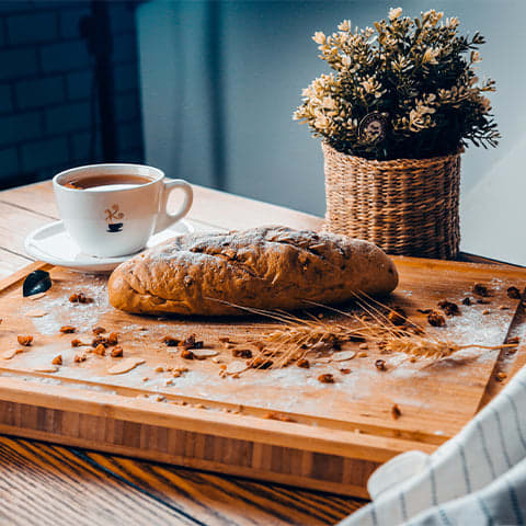 A small loaf of bread sitting on a small cutting board in front of a small plant and a small cup of coffee.